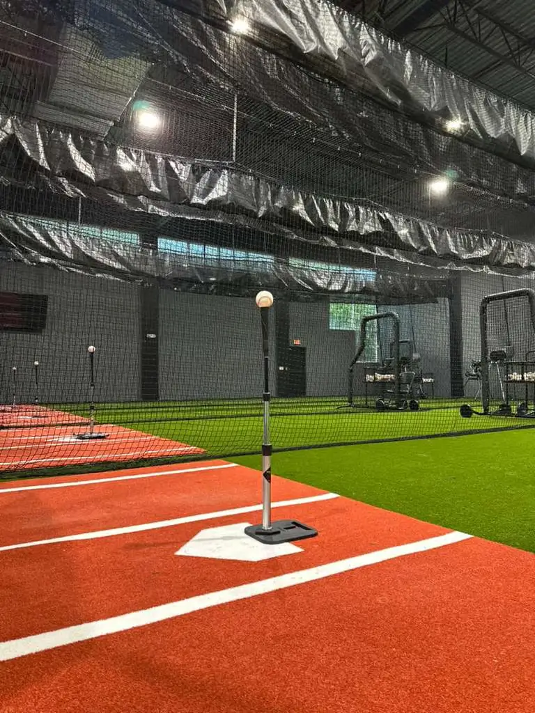 Professional indoor baseball training facility featuring red and green turf, batting tees with baseballs, and protective black netting.